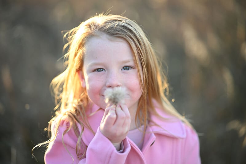 A young girl with long blonde hair and a pink coat holds a dandelion puff up to her face, smiling softly, outdoors with a blurred natural background.