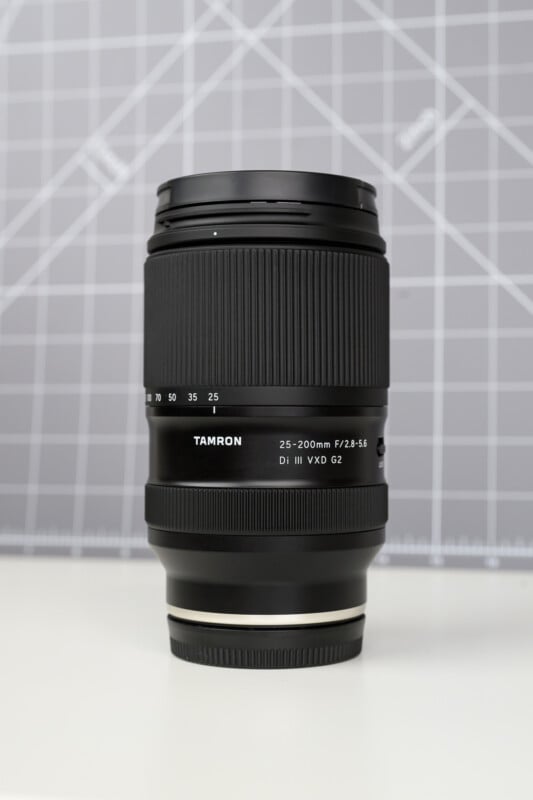 A Tamron 25-200mm f/2.8-5.6 Di III VXD G2 camera lens stands upright on a white surface with a gray grid-patterned background.