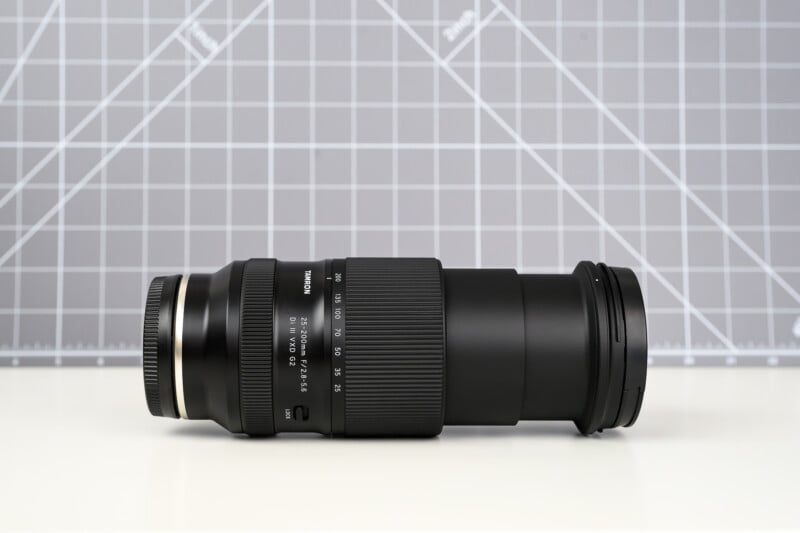 A black camera zoom lens is placed horizontally on a white surface, with a gray grid background behind it. The lens shows detailed markings and a textured grip.