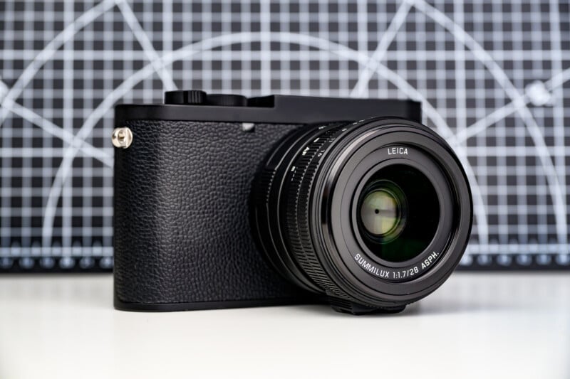 Cámara digital Leica negra con lente grande sobre superficie blanca frente a un fondo de cuadrícula blanco y negro con marcas circulares.