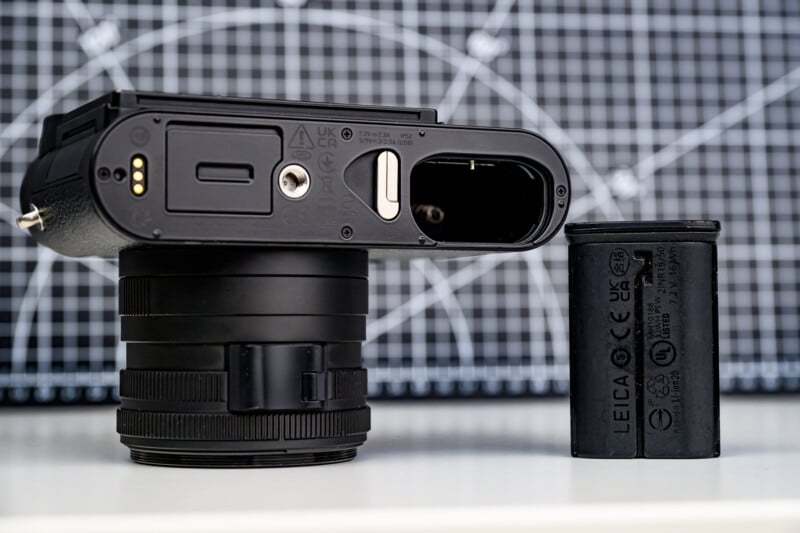 Primer plano de la cámara tumbada de lado, la caja de la batería abierta, la batería extraída y colocada al lado de la cámara. Detrás de ellos se puede ver un fondo cuadriculado con líneas blancas.