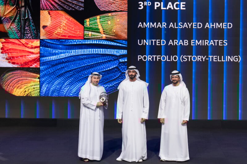 Three men in traditional Emirati attire stand on stage; one holds a trophy. Behind them, colorful abstract images are displayed along with text announcing "3rd Place" in portfolio storytelling for Ammar Alsayed Ahmed from the United Arab Emirates.