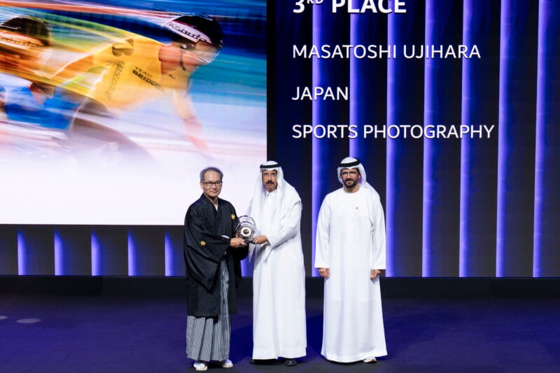 Three men stand on a stage during an award ceremony. The man on the left wears traditional Japanese attire and holds a trophy. The two men on the right wear white kanduras. A large display in the background shows text and a sports photo.
