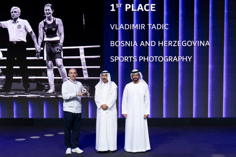 Three men stand on stage holding a trophy. Behind them, a large screen shows a boxing photo and the text: "1st Place Vladimir Tadic Bosnia and Herzegovina Sports Photography.