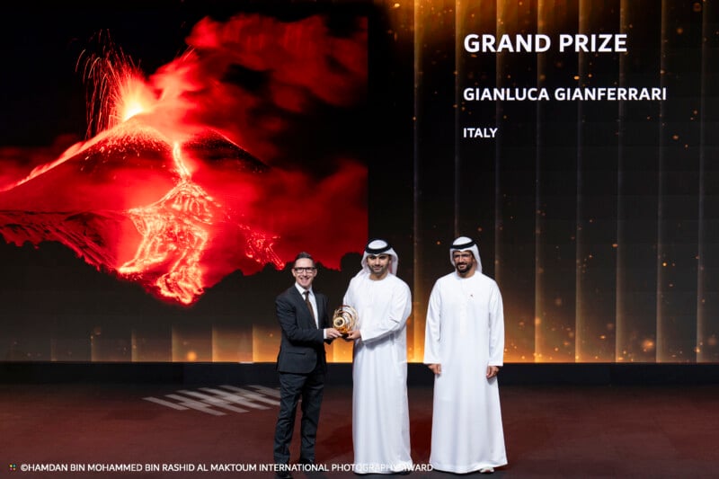 Tres hombres suben al escenario de la ceremonia de premiación; uno sostiene un trofeo. Detrás de ellos hay una foto de un volcán en erupción con las palabras 
