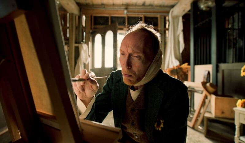 A man with a bandaged head intently paints on a canvas in a sunlit, rustic studio. He wears a dark coat with a flower on the lapel, and light streams through windows behind him.