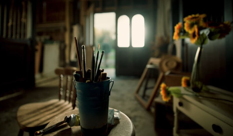 A sunlit artist's studio with paintbrushes in a cup on a table, a vase of yellow flowers, an easel, and chairs. The room is rustic, with soft light streaming through the open door in the background.