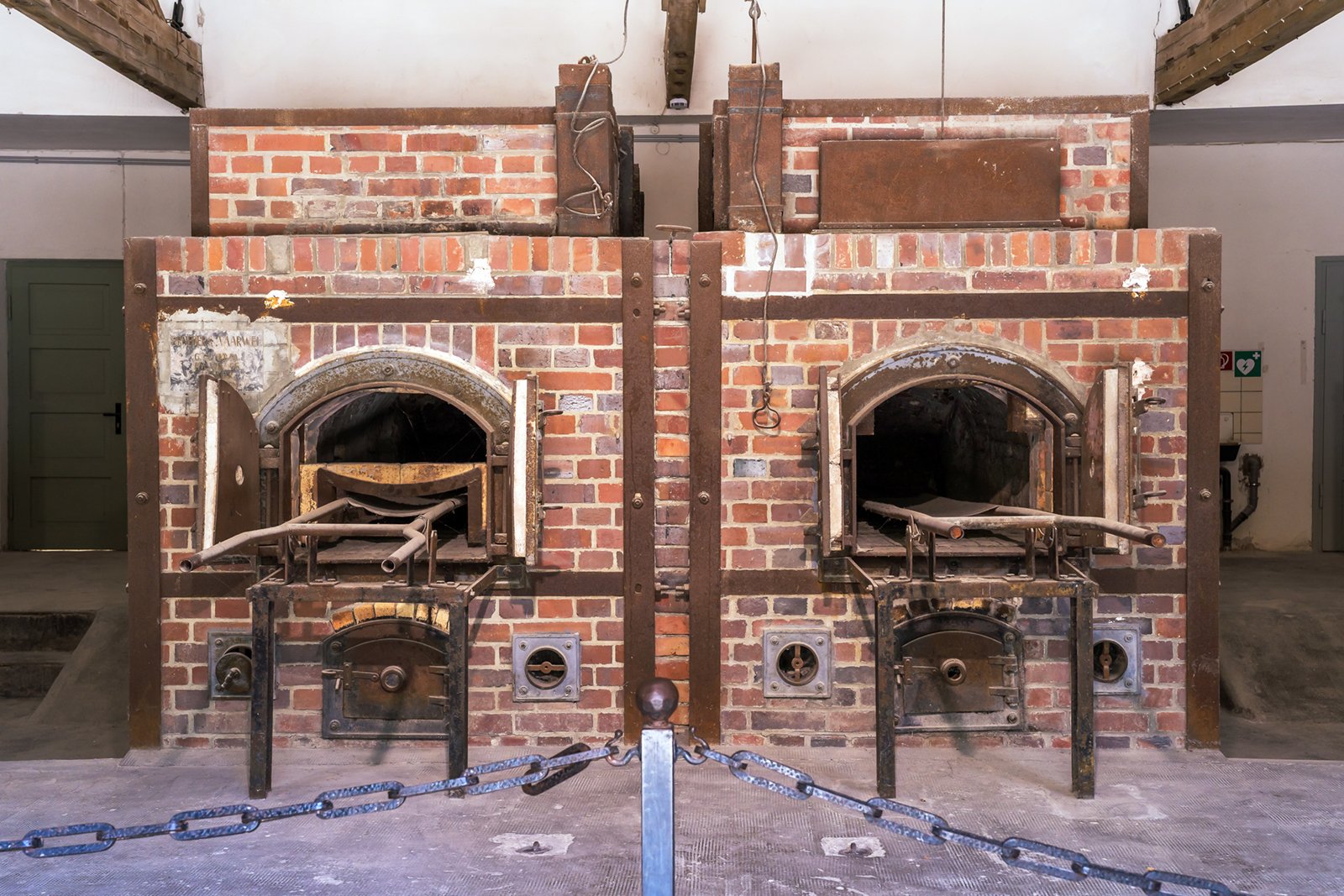 Two brick crematorium ovens with open metal doors and racks inside, situated side by side in an empty, industrial room. A metal chain barrier is positioned in front of the ovens.