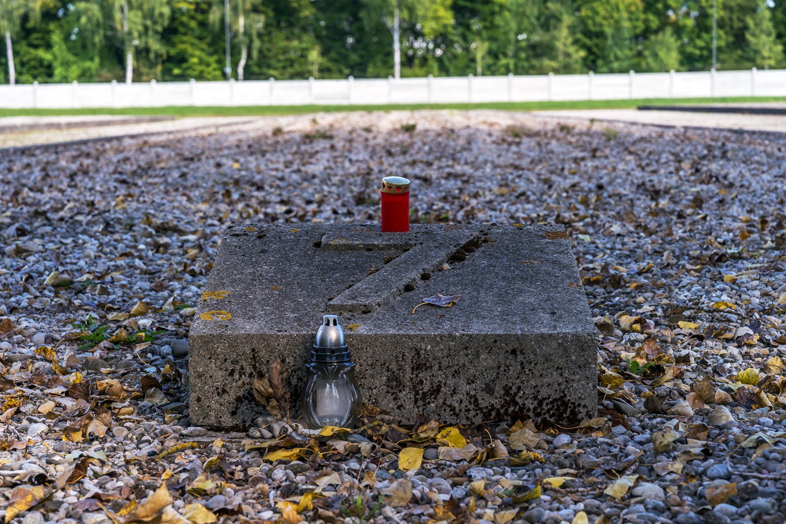 A concrete slab with metal numbers on it sits on gravel, surrounded by fallen leaves. A red candle rests on top, and a white lamp is placed in front. In the background, there's a green lawn, trees, and a white wall.