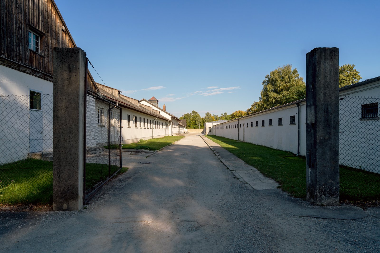 A paved pathway flanked by two tall concrete pillars leads between long, white buildings with barred windows and fenced yards under a clear blue sky, with trees visible in the background.