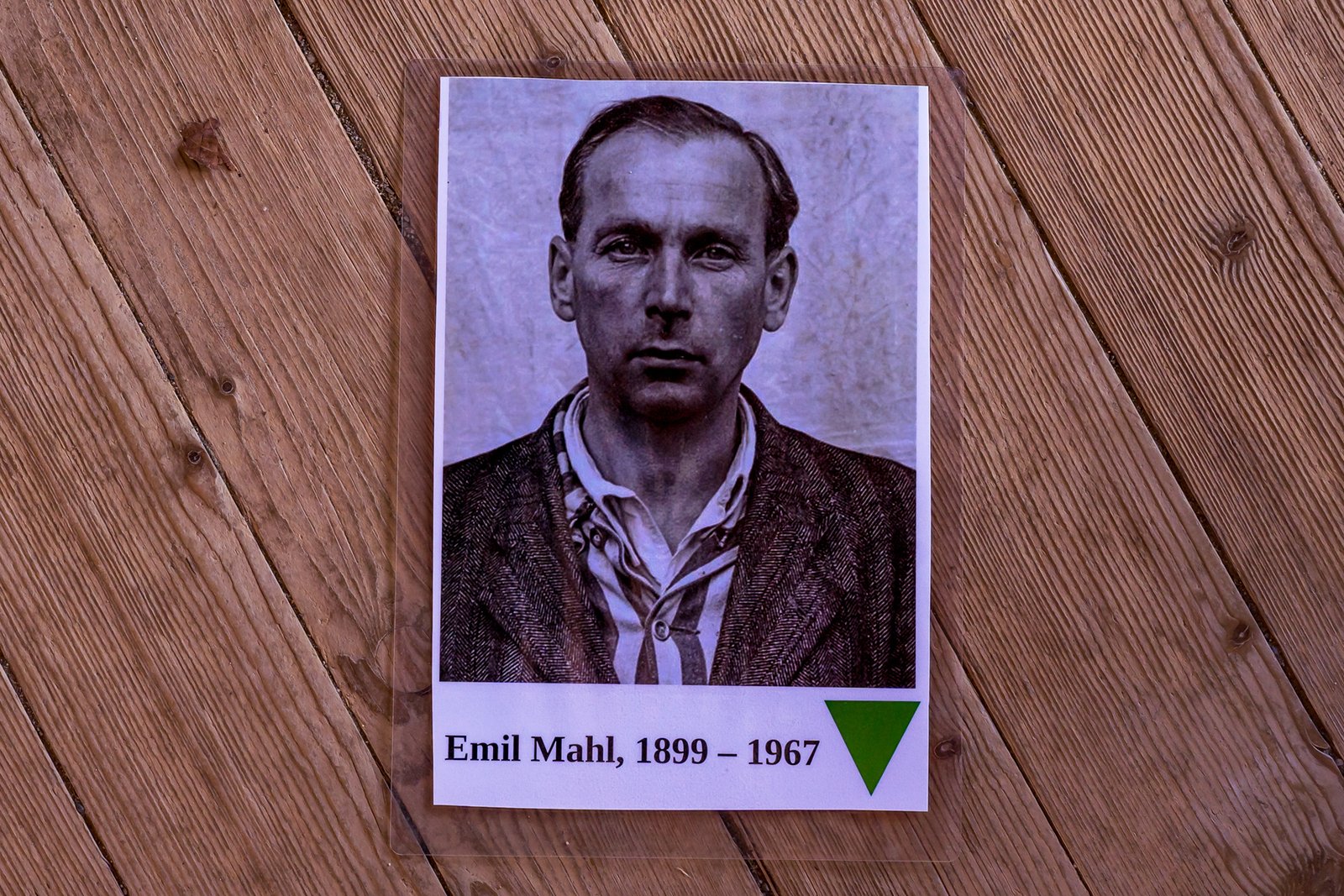 A black and white photo of Emil Mahl with text below reading "Emil Mahl, 1899 – 1967" and a green triangle, placed on a wooden floor with diagonal planks.
