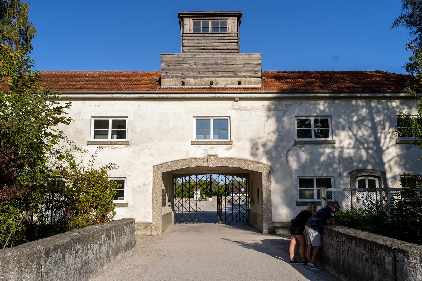 Two people stand near the entrance gate of a historic building with a red roof and central tower, surrounded by greenery, under a clear blue sky.