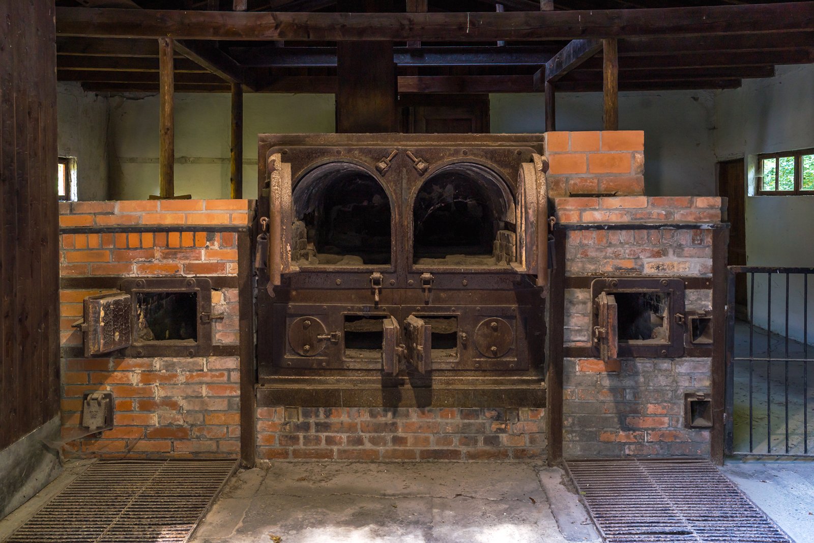 Historic brick crematorium oven with iron doors and mechanisms, located inside an old building with exposed wooden beams and barred windows, illuminated by natural light.