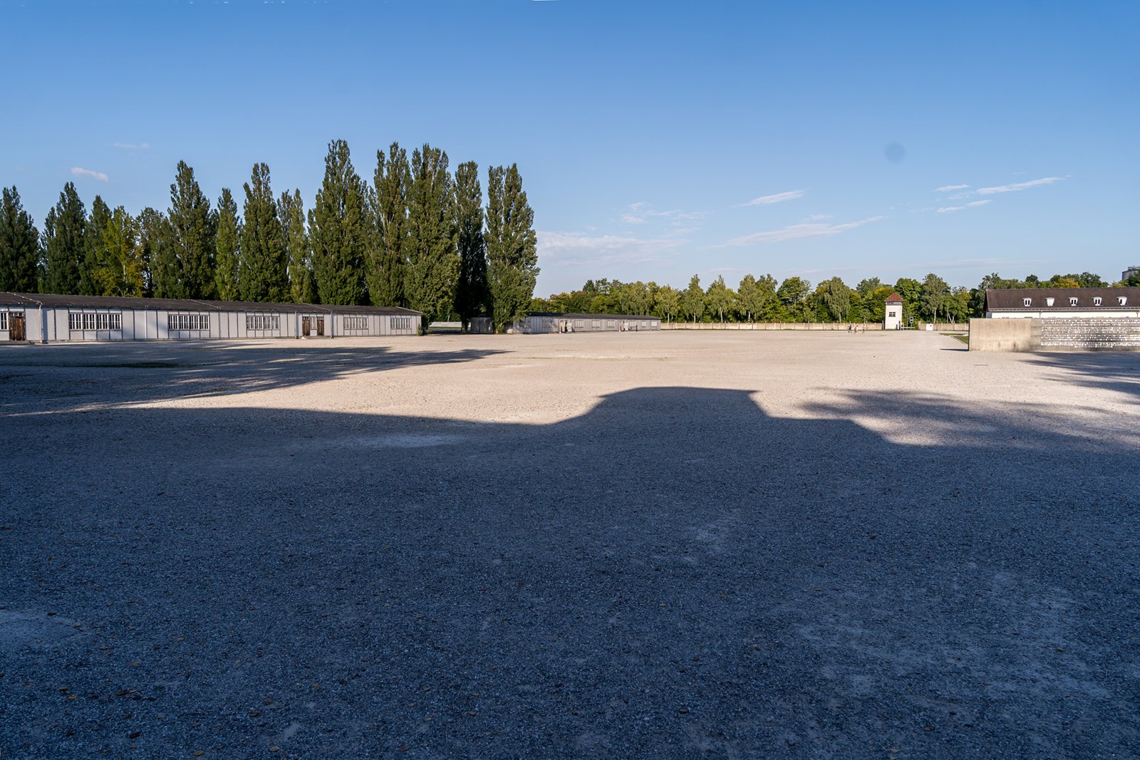 A wide, empty gravel courtyard under a clear blue sky, bordered by trees on the left and low buildings along the edges, with long shadows cast across the ground.