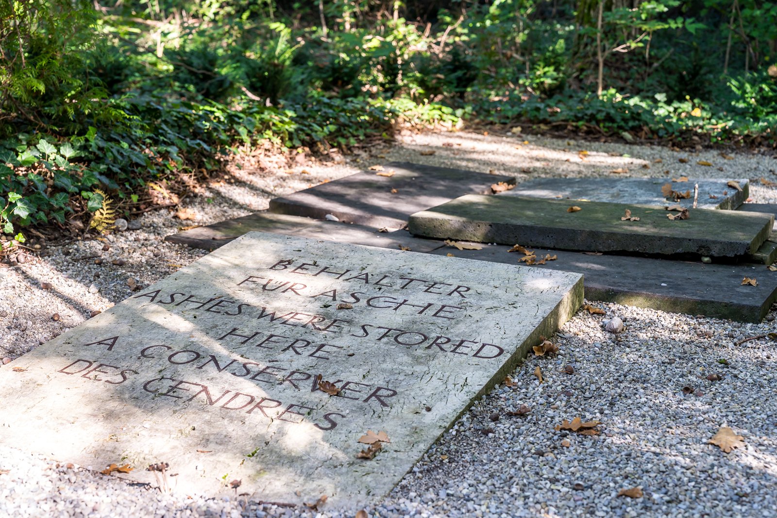 A stone plaque in a wooded area reads, "BEHÄLTER FÜR ASCHE ASHES WERE STORED HERE A CONSERVER DES CENDRES," surrounded by gravel, fallen leaves, and greenery.