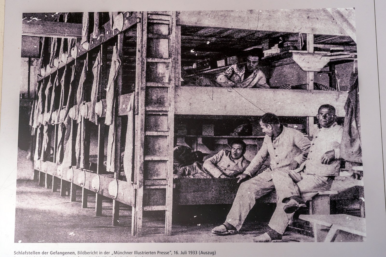 Black and white photo showing several men in prison uniforms sitting and lying on wooden bunk beds in a crowded dormitory. Some look toward the camera, while others rest or talk. The scene appears somber and confined.