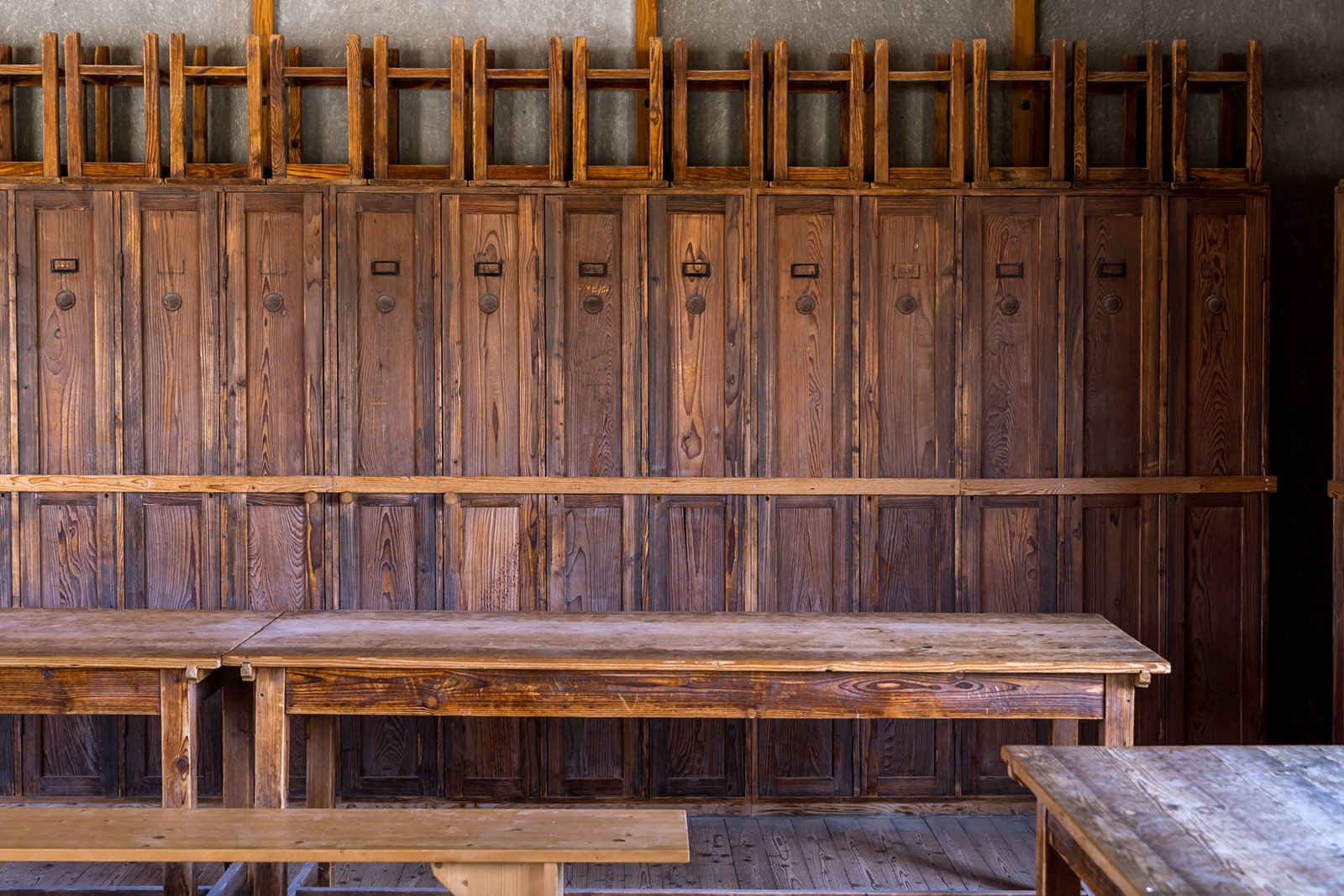 Rows of vintage wooden lockers with nameplates line the back wall of an old classroom; wooden benches and tables are in the foreground, and sunlight casts soft shadows across the scene.