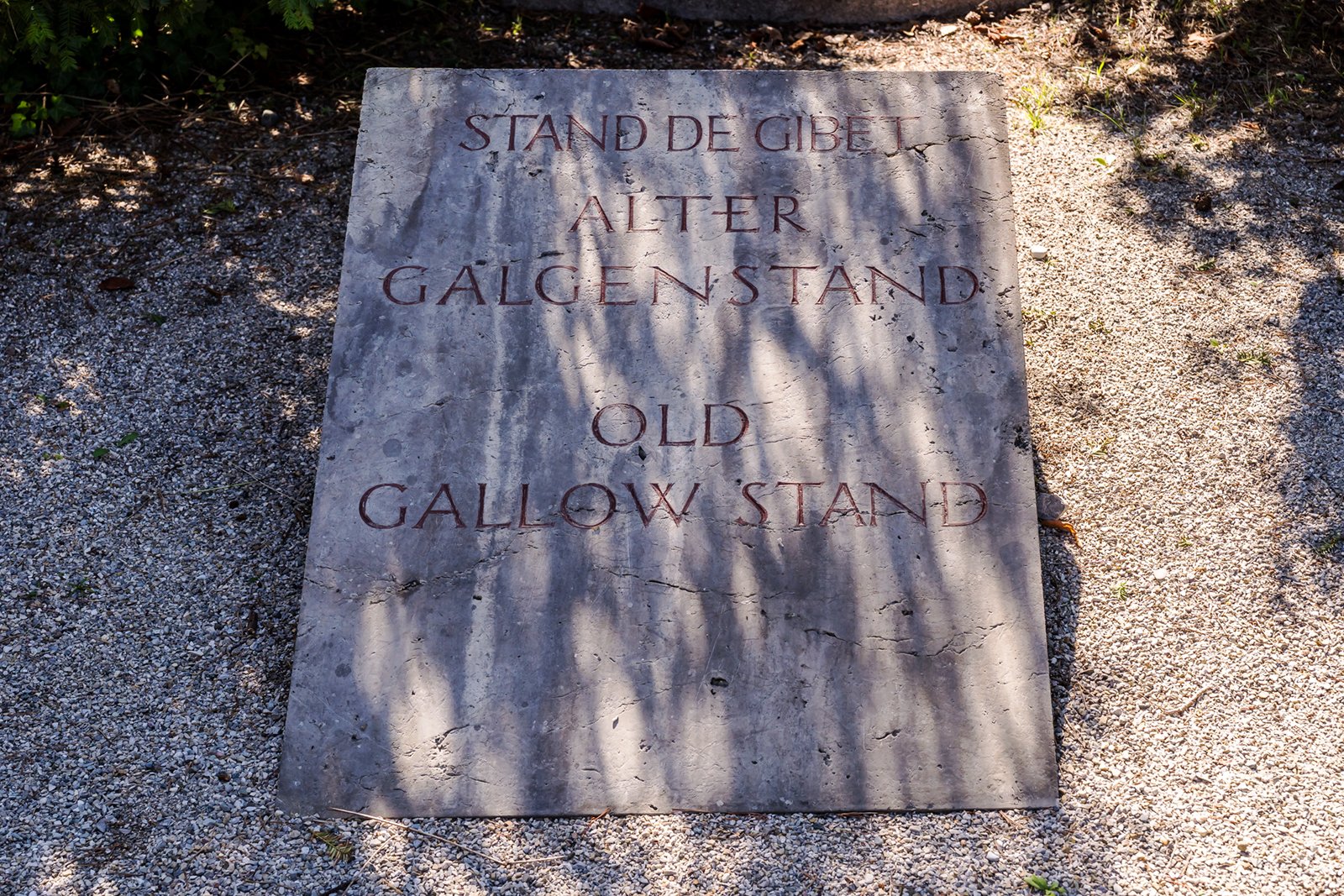 A stone plaque set in gravel displays text in German and English: "STAND DE GIBELT, ALTER GALGENSTAND, OLD GALLOW STAND." Shadows of nearby plants fall across the plaque.