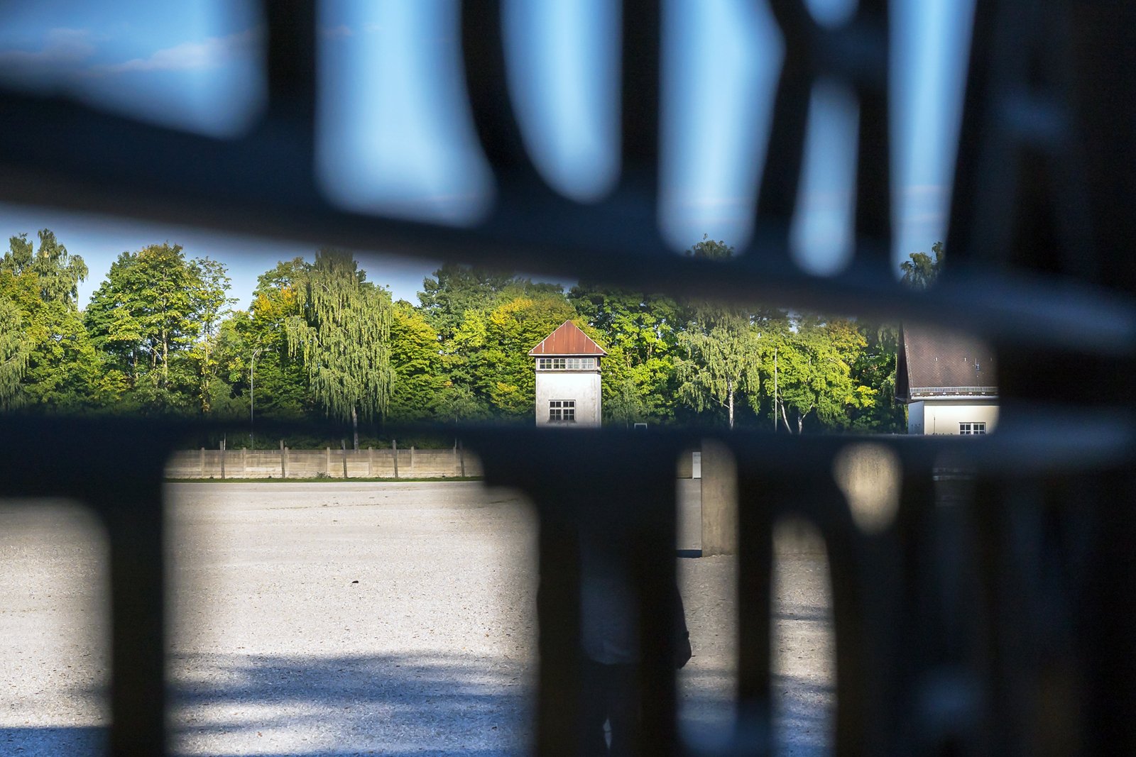 View through a metal gate with bars, looking out onto an open courtyard with a building in the distance, surrounded by trees under a partly cloudy sky.