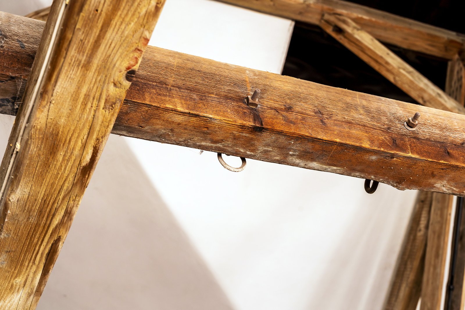 Close-up of wooden beams forming part of a rustic structure, with several metal rings and nails attached to the wood; a white surface is visible in the background.