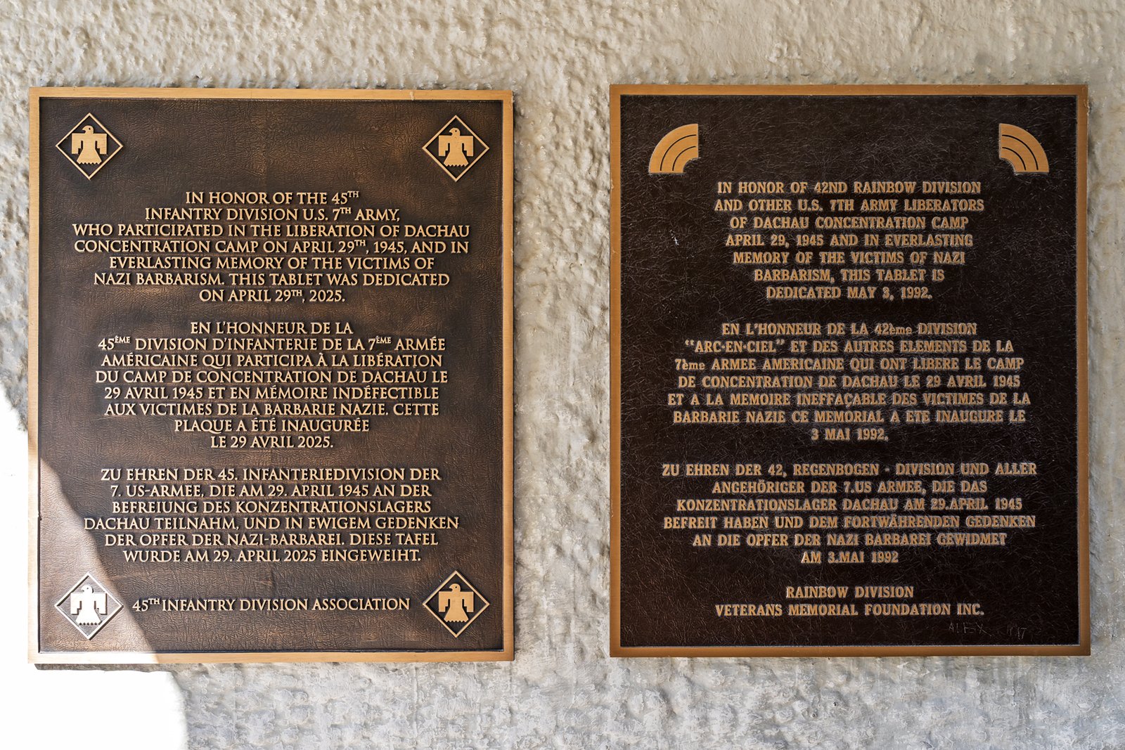 Two commemorative plaques on a wall honor U.S. Army divisions for liberating Dachau concentration camp in April 1945, with inscriptions in English, French, and German. Each plaque features military insignias.