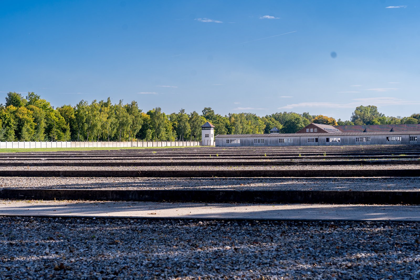 Wide view of Dachau concentration camp memorial site with rows of gravel, a guard tower, and buildings in the distance, surrounded by trees under a clear blue sky.
