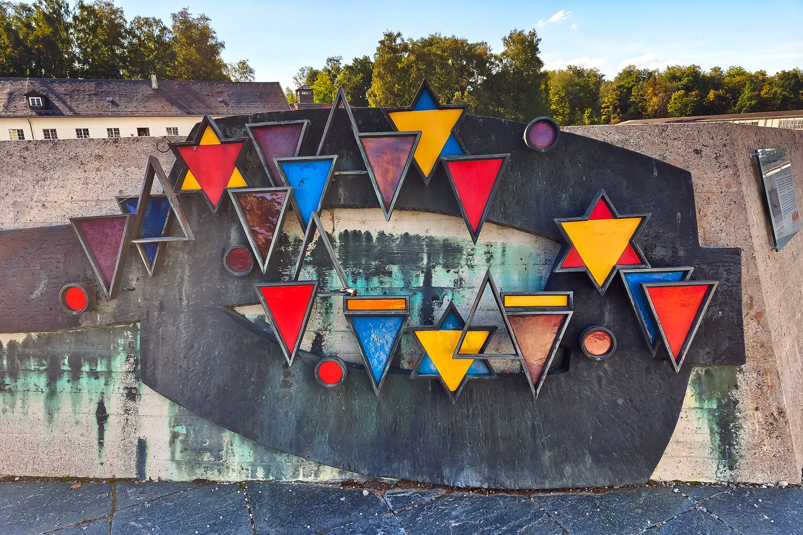 A colorful outdoor abstract sculpture features red, yellow, and blue triangles and stars arranged on a concrete wall, with trees and a building in the background.
