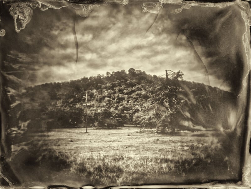 Fotografía de estilo vintage en tonos sepia de una pradera con un árbol y una colina boscosa al fondo rodeada por un borde desgastado, rayado y texturizado. La escena parece antigua y atmosférica.