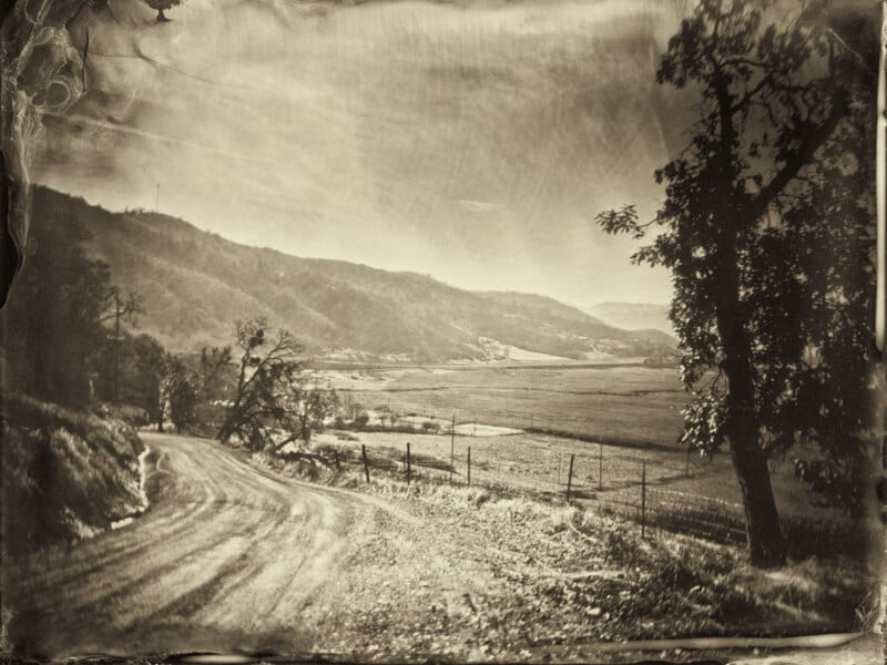 Fotografía en tonos sepia de un sinuoso camino de tierra que serpentea a través de un paisaje rural, rodeado de colinas y campos, con árboles que enmarcan la escena y montañas distantes al fondo.