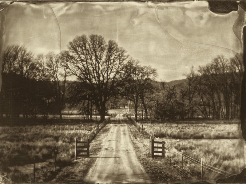 Una fotografía de estilo retro en tonos sepia de un camino rural de tierra flanqueado por puertas y vallas de madera, que conduce a árboles desnudos y colinas bajo un cielo nublado en la distancia.