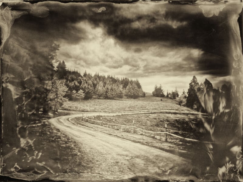 Foto en tonos sepia de un sinuoso camino de tierra que serpentea a través de un prado con árboles en una ladera bajo un cielo nublado, que se asemeja a una imagen antigua de estilo vintage o de tabla mojada.