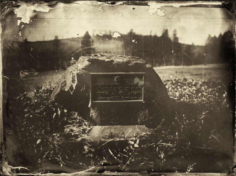 Una fotografía en tonos sepia de una placa conmemorativa montada sobre una gran roca y situada en un prado, con árboles y colinas visibles en el fondo borroso; la imagen parece vieja y desgastada.