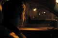 A person sits in a car at night, looking out the passenger-side window toward a dimly lit street with distant streetlights and a shadowy figure on the sidewalk.