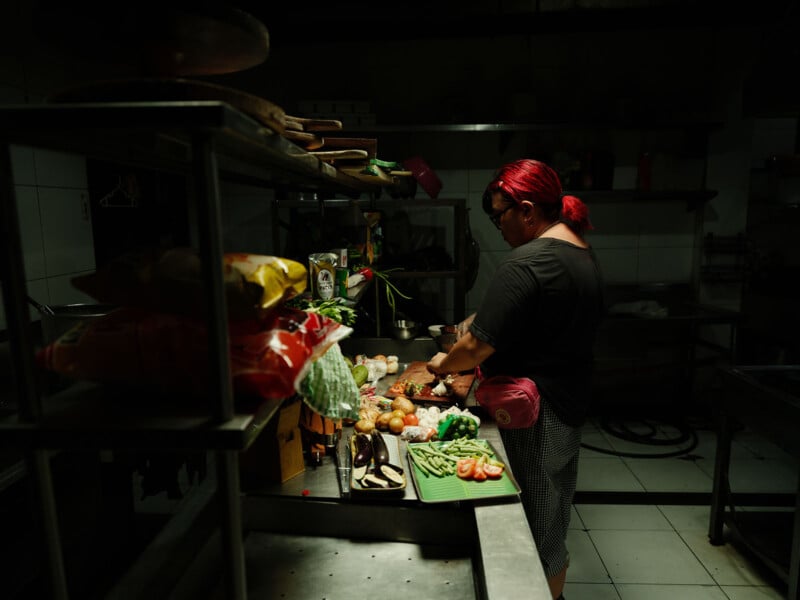 En una cocina con poca luz, un hombre pelirrojo prepara verduras frente a un mostrador desordenado rodeado de diversos ingredientes y utensilios de cocina.