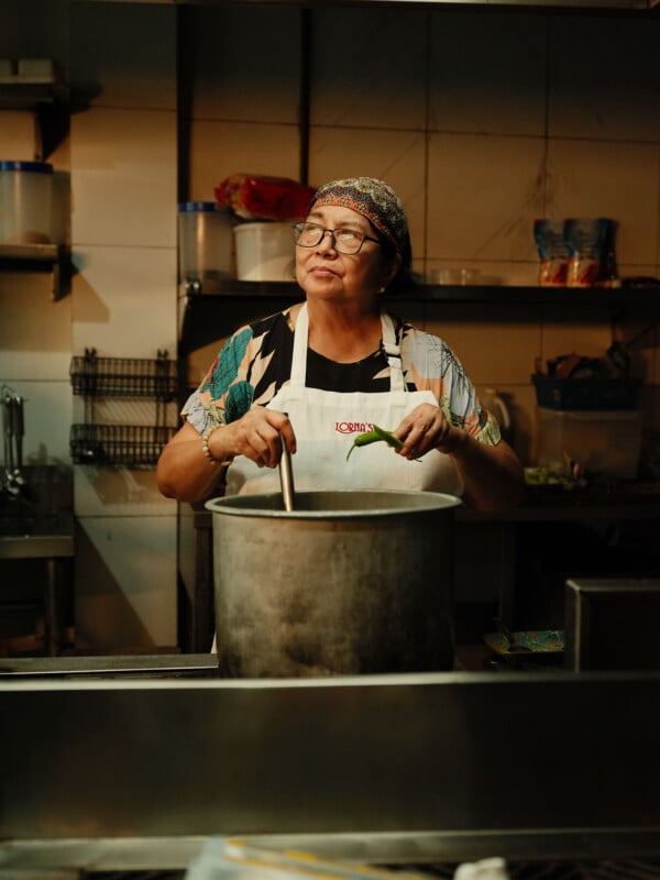 Una mujer con gafas, un pañuelo estampado en la cabeza y un delantal remueve comida en una olla grande en la cocina. Al fondo se ven estantes con recipientes e ingredientes.