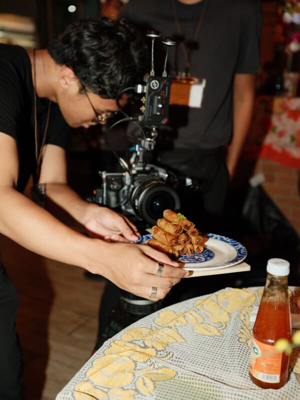 Un hombre coloca con cuidado un plato de rollitos de primavera frente a una cámara profesional, preparándose para fotografiar la comida. Una botella de salsa se encuentra sobre la mesa con un mantel floral amarillo en primer plano.