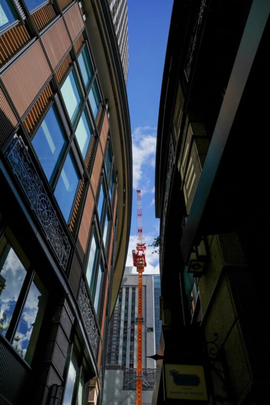 A view looking up between two tall buildings, revealing a red construction crane and a skyscraper in the background against a blue sky with scattered clouds.