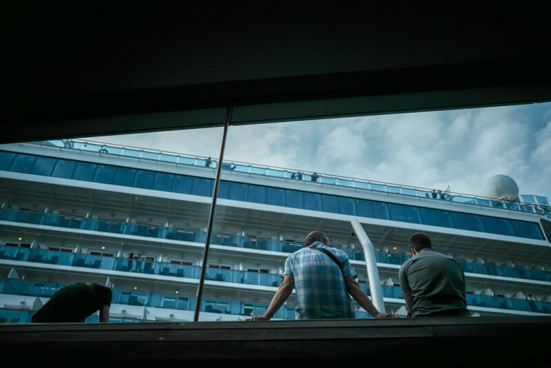 Three people are seen from behind, looking out at a large cruise ship with multiple decks and balconies under a cloudy sky. The photo is taken through a window, framing the scene from a low angle.