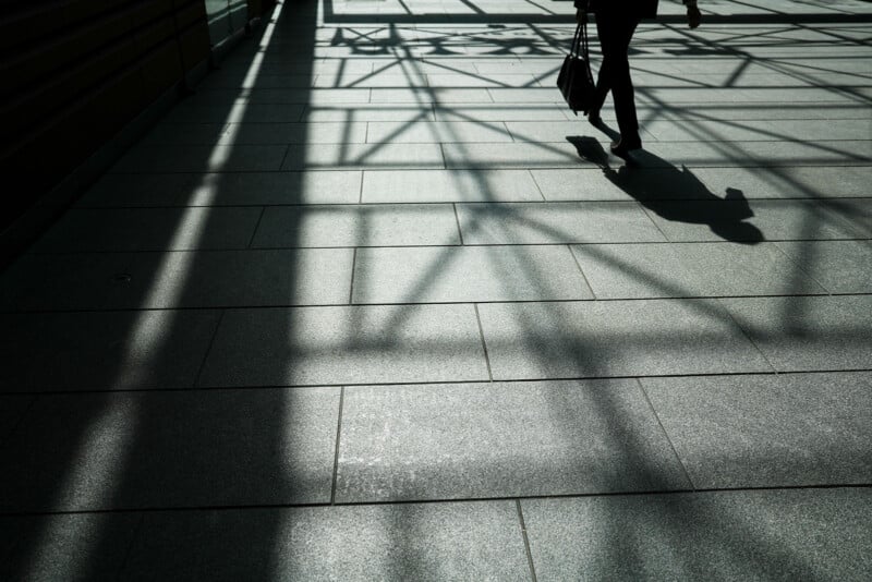 A person walks inside a building, casting a shadow on the tiled floor, with strong geometric shadows from the overhead structure creating a pattern across the ground.