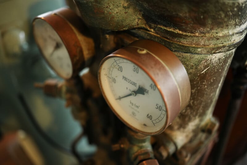 Close-up of two old, round pressure gauges mounted on a weathered metallic surface, showing numerical readings and labeled "PRESSURE." The gauges appear to be part of industrial machinery.
