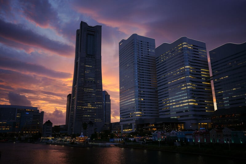 Tall modern skyscrapers are silhouetted against a dramatic sunset sky with vibrant purple, orange, and pink clouds, reflecting on the water in a cityscape scene.