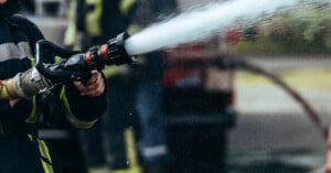 A firefighter wearing protective gear sprays water from a hose, with water forcefully streaming out. The background is blurred, showing other firefighters and equipment.