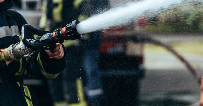 A firefighter wearing protective gear sprays water from a hose, with water forcefully streaming out. The background is blurred, showing other firefighters and equipment.