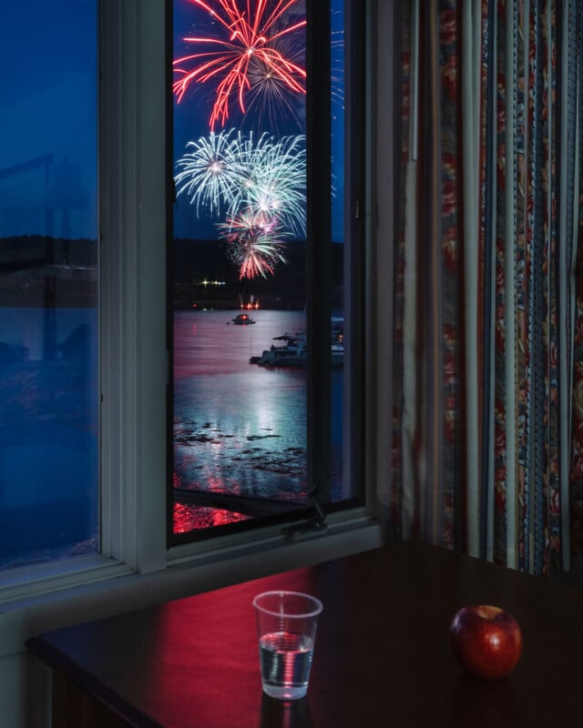 Había una mesa en la habitación con un vaso de plástico transparente con agua y una manzana roja; A través de una ventana abierta, coloridos fuegos artificiales iluminaban el cielo nocturno sobre el agua donde estaban amarrados los barcos.