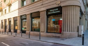 A corner view of a Fujifilm House of Photography store with large windows, black awnings, and a stone facade on a quiet city street. Bollards line the sidewalk in front of the shop.