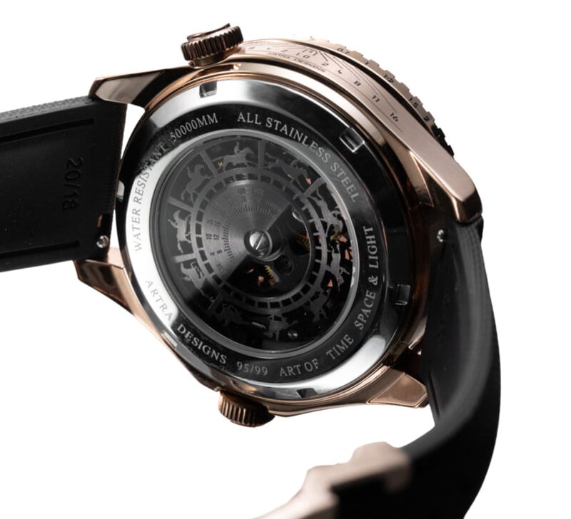 Primer plano de la parte trasera de un reloj de lujo con caja transparente que revela engranajes complejos. El texto grabado es el siguiente. "Todo de acero inoxidable," "El arte del tiempo, el espacio y la luz," y "Diseño ATRA 95/99.