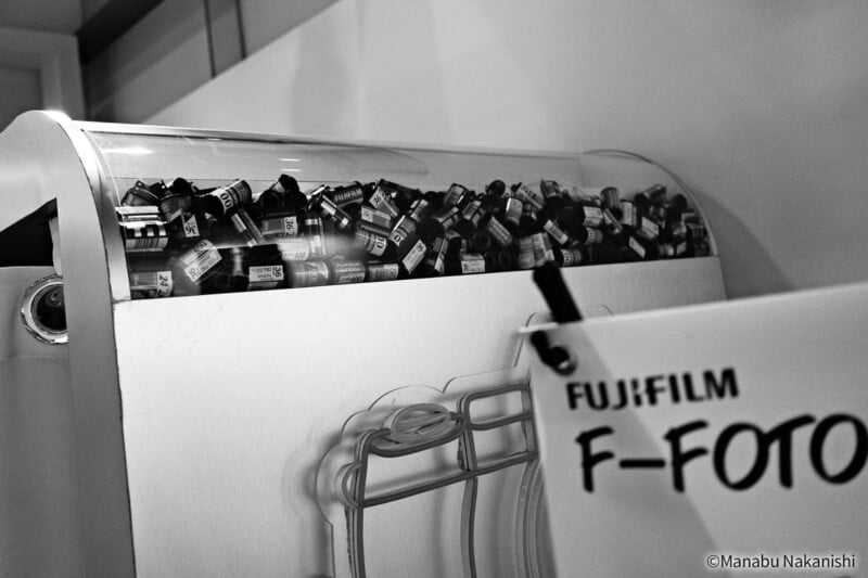 En el mostrador de la tienda había un contenedor lleno de rollos de película de 35 mm. Hay un logotipo de FUJIFILM F-FOTO parcialmente visible en primer plano. La imagen está en blanco y negro.