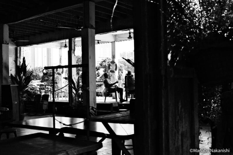 Fotografía en blanco y negro de una terraza exterior cubierta con mesa y sillas. Dos personas se sientan y conversan al fondo, rodeadas de pilares, plantas y edificios de la ciudad visibles a lo lejos.