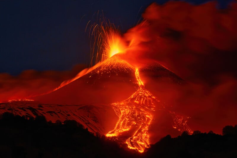 Un volcán entró en erupción por la noche, con lava de color naranja brillante fluyendo por sus costados y chispas disparadas hacia el cielo oscuro, rodeadas de humo y cenizas.