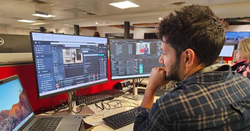 Un hombre sentado en su escritorio en una oficina y mira dos grandes monitores de computadora que muestran software de edición de video. Al fondo, otra persona trabaja en una estación de trabajo cercana.